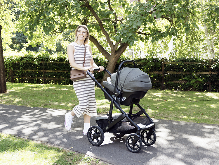 Mother walking with gray Joie Vinca pushchair and Sprint infant car seat attached, in outdoor park.
