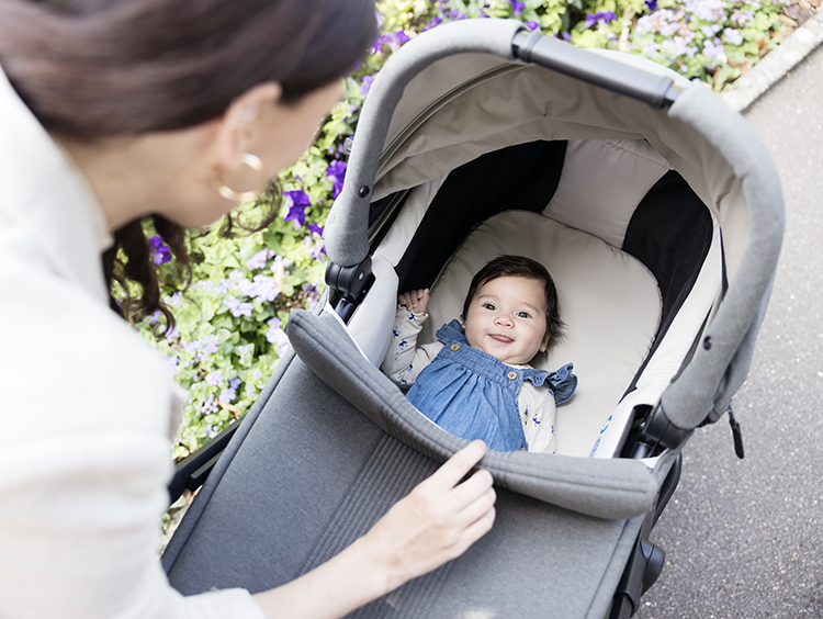 Mom smiling down at baby in carry cot on Joie Signature vinca pram beside flowerbed of purple flowers.
