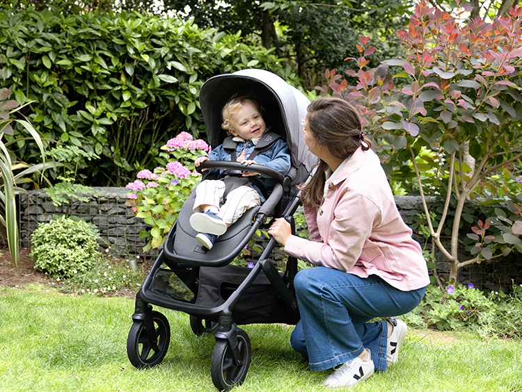Mother smiling at toddler in a gray Joie Vinca pushchair in outdoor garden.
