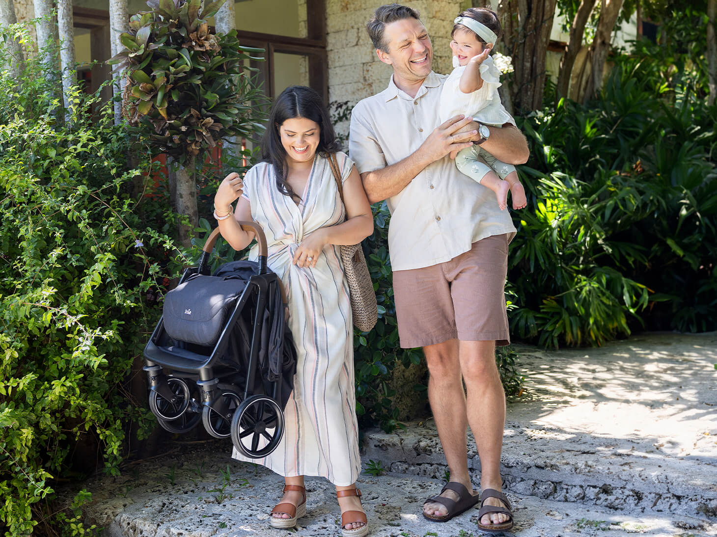 mom holding folded nutmeg stroller in shale with dad and baby in garden.