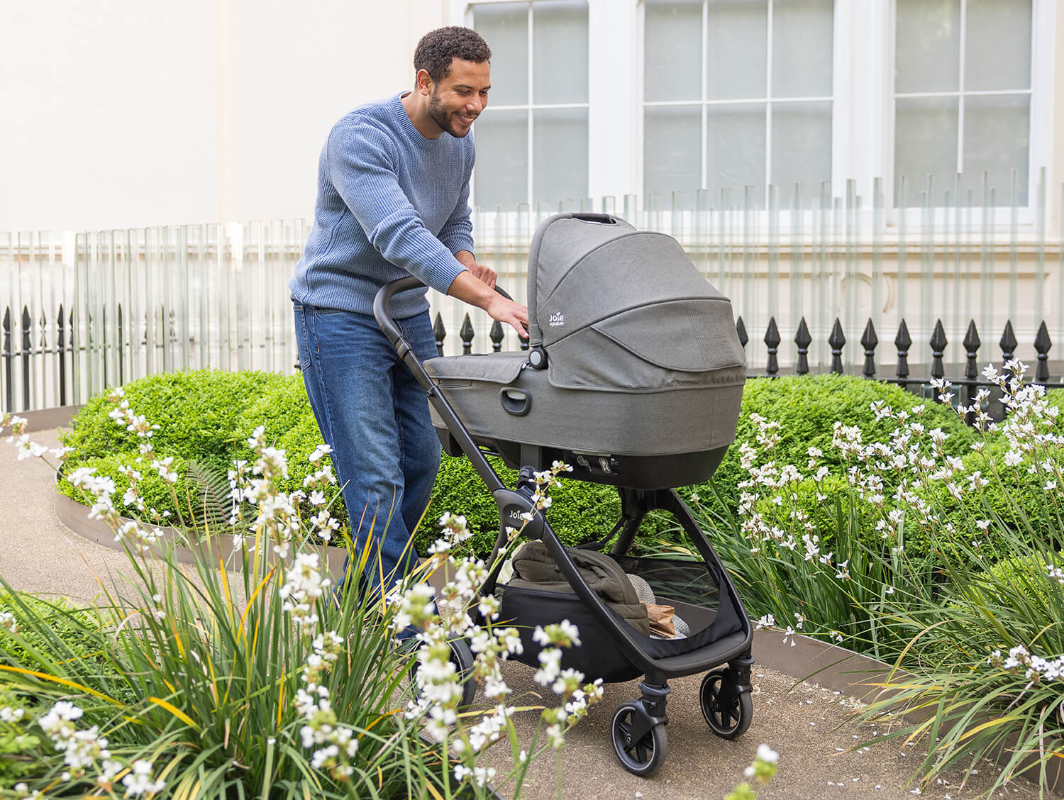Dad walking through a garden while pushing the Joie valora pram with a carry cot attached and interacting with his baby