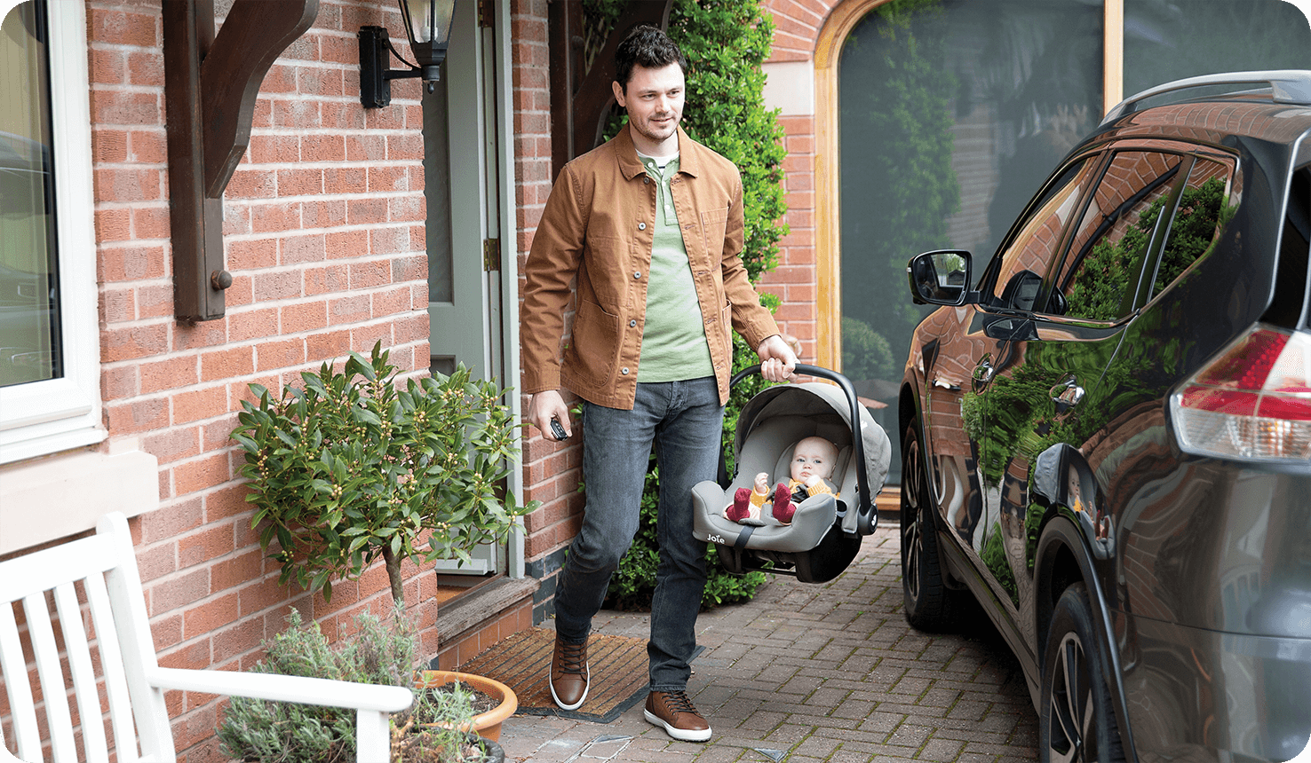 dad walking out of his house going towards his car carrying the joie i-snug 2 infant car seat with his baby in it
