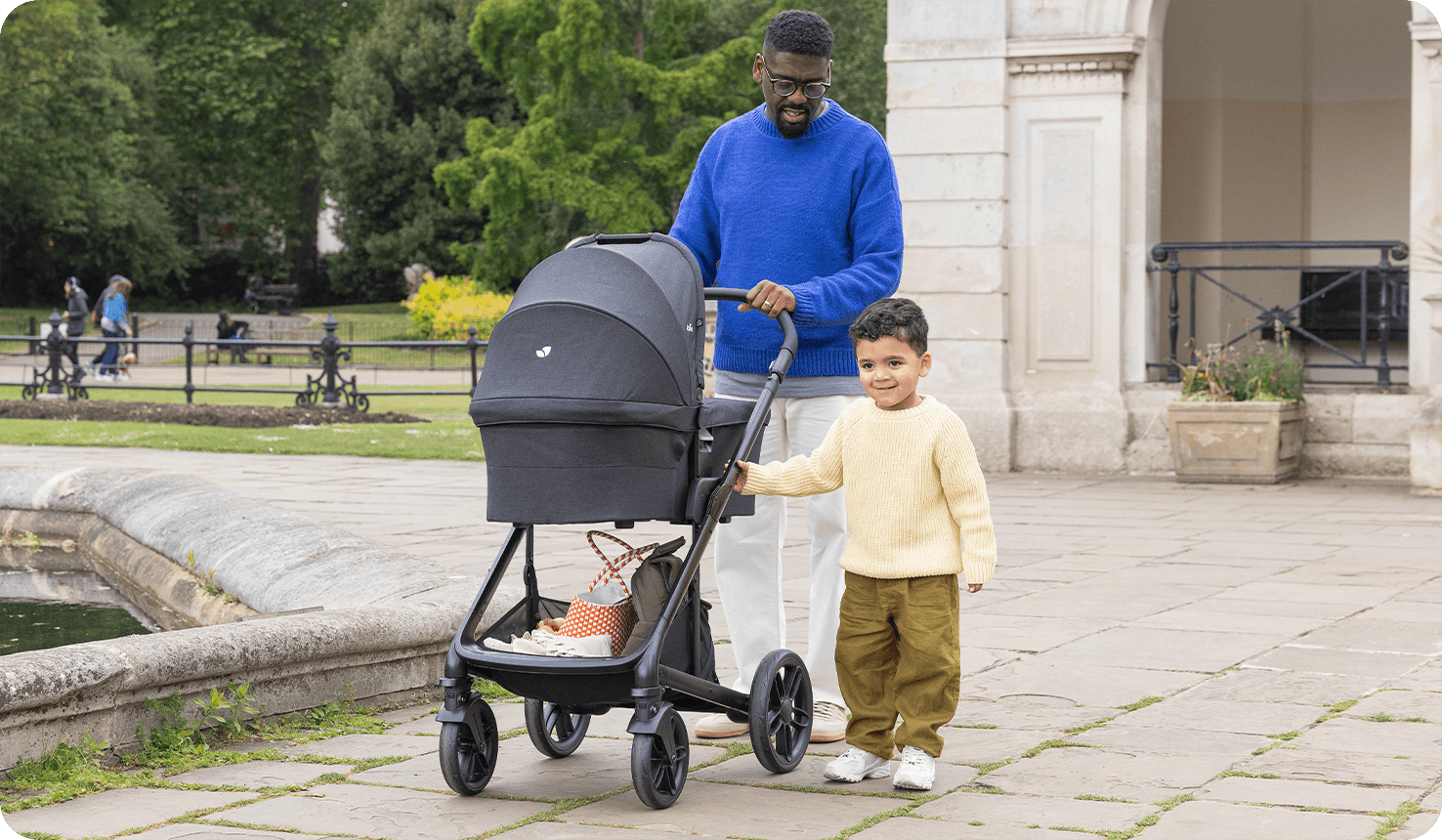 Dad and his younger son walking in a park and pushing the Joie chrome 2 pram with the carry cot attached