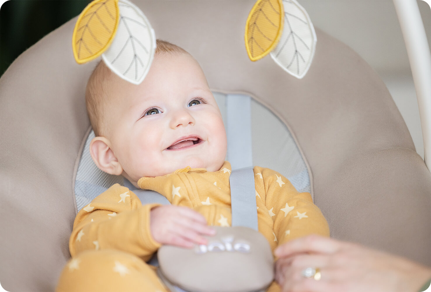 Bébé souriant dans la balancelle Joie whimsy, regardant les 2 jouets suspendus à l’arche de jeu fournie