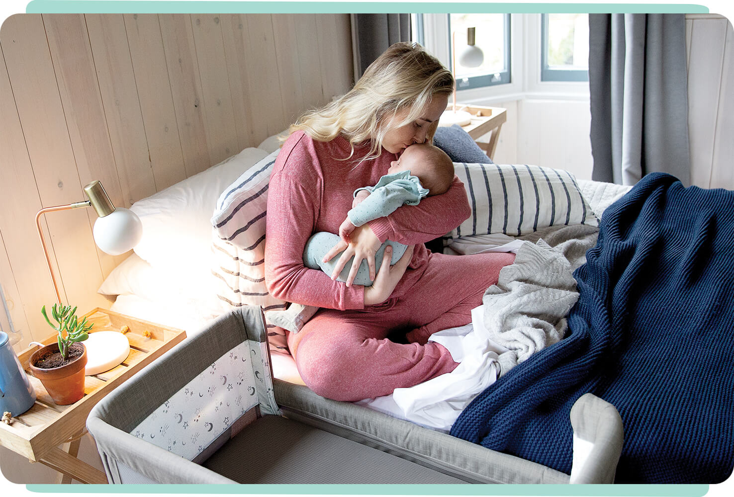  Mom kissing baby while sitting in bed next to light grey Joie roomie glide bedside crib.