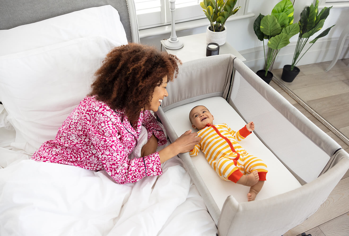 Die Mutter liegt im Bett und interagiert mit dem Baby, das im Roomie Go Kinderbett/Reisebett liegt.