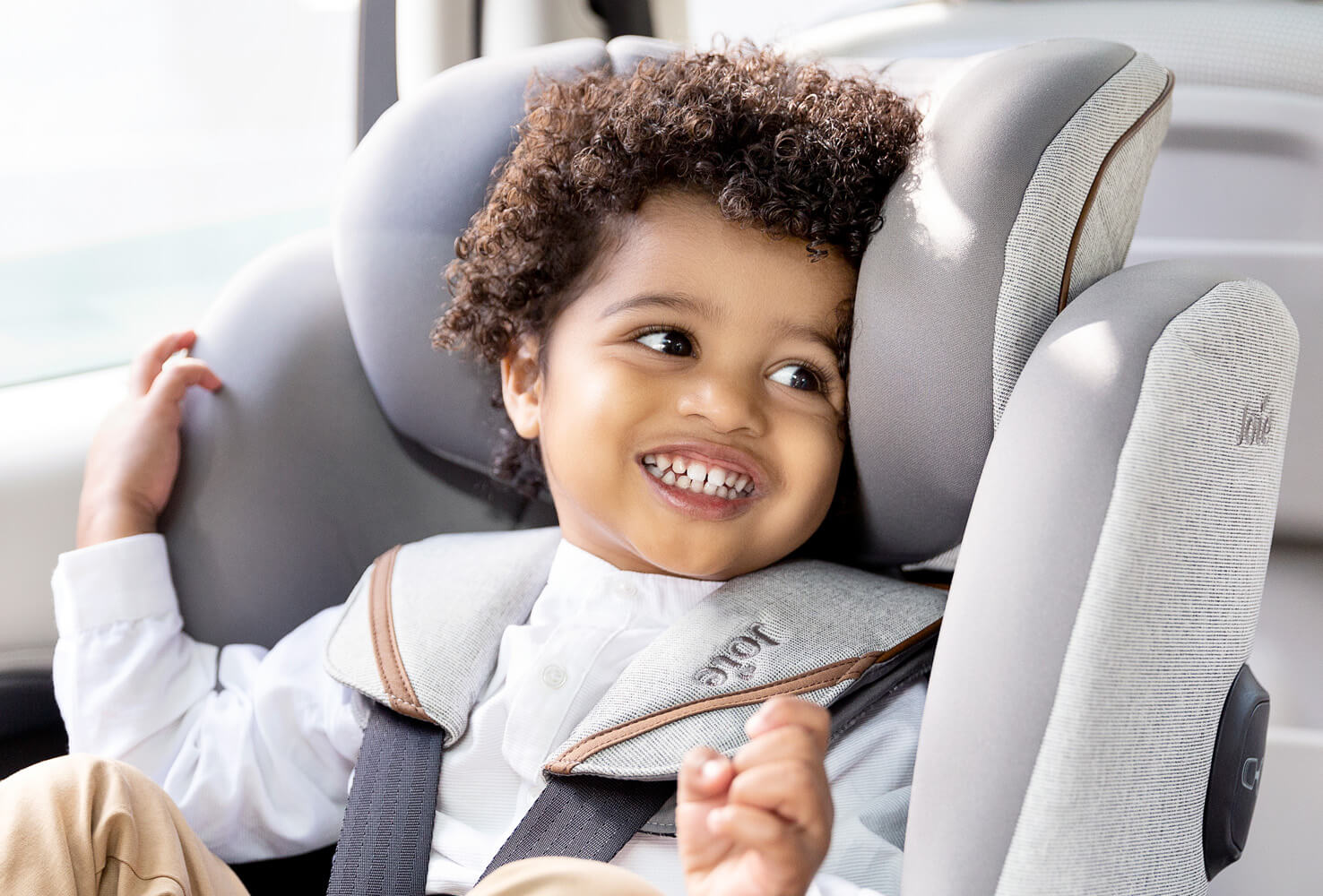  A young boy with curly brown hair seated in a gray Joie I-Quest car seat, smiling at something off camera.