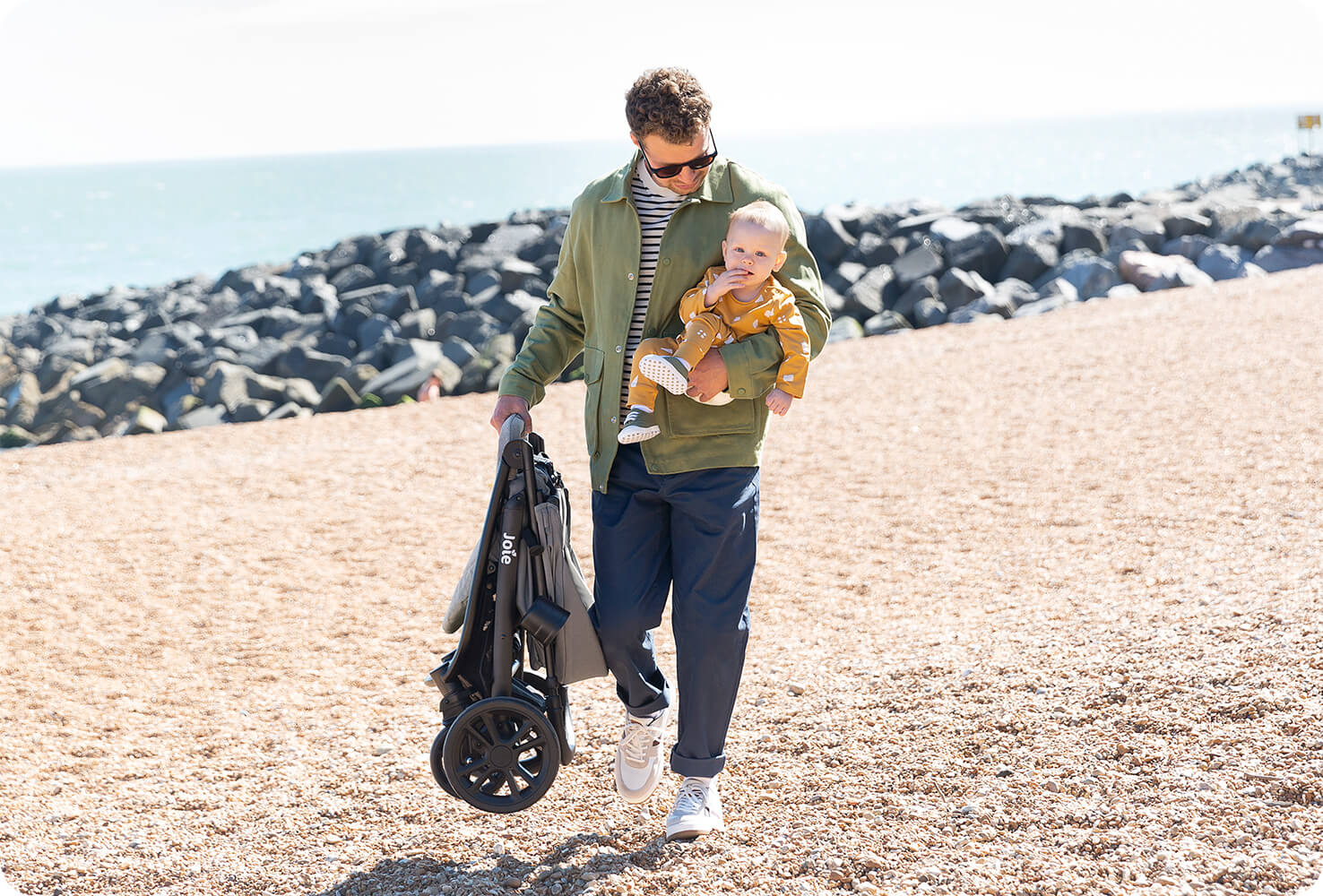 Ein Vater geht am Strand spazieren und trägt ein Baby auf einem Arm und einen zusammengeklappten litetrax 4 dlx Kinderwagen in der anderen Hand.