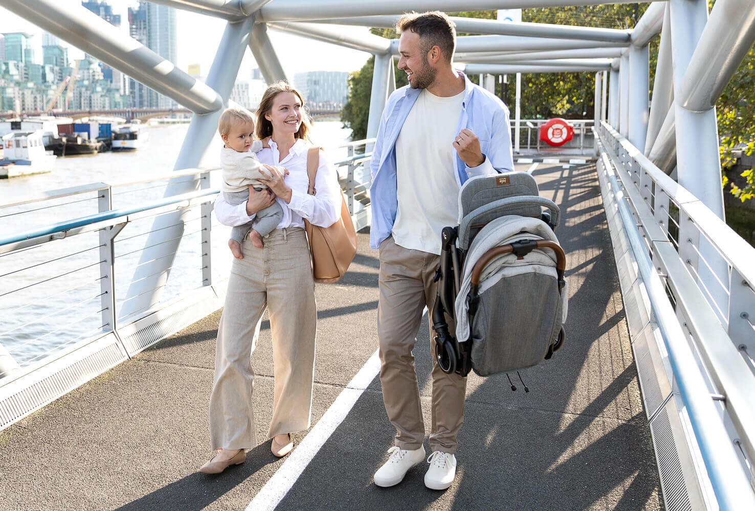 Poussette Joie tourist repliée, portée par un père pendant que la mère et son bébé marchent à ses côtés sur un pont.