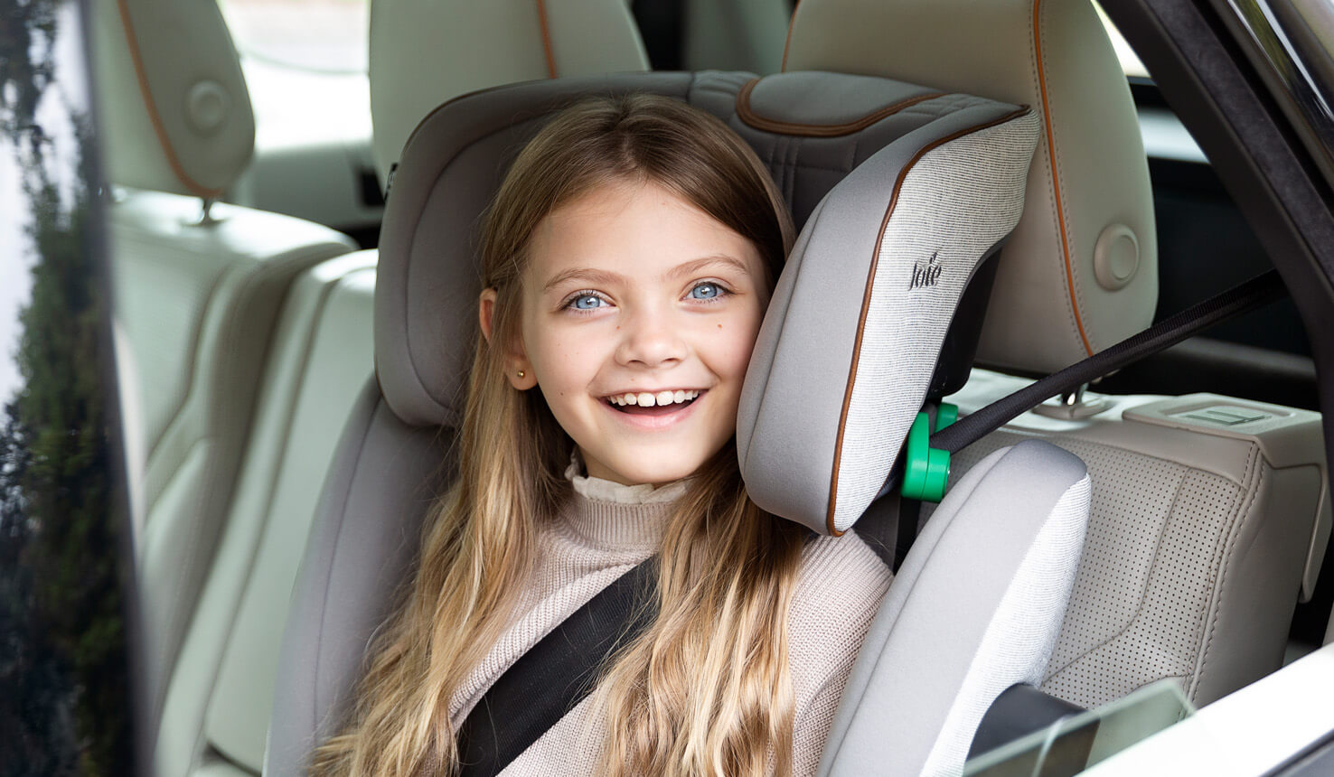 Junges Mädchen lächelt aus dem Autofenster, während es in einem Joie i-Traver Autokindersitz sitzt.