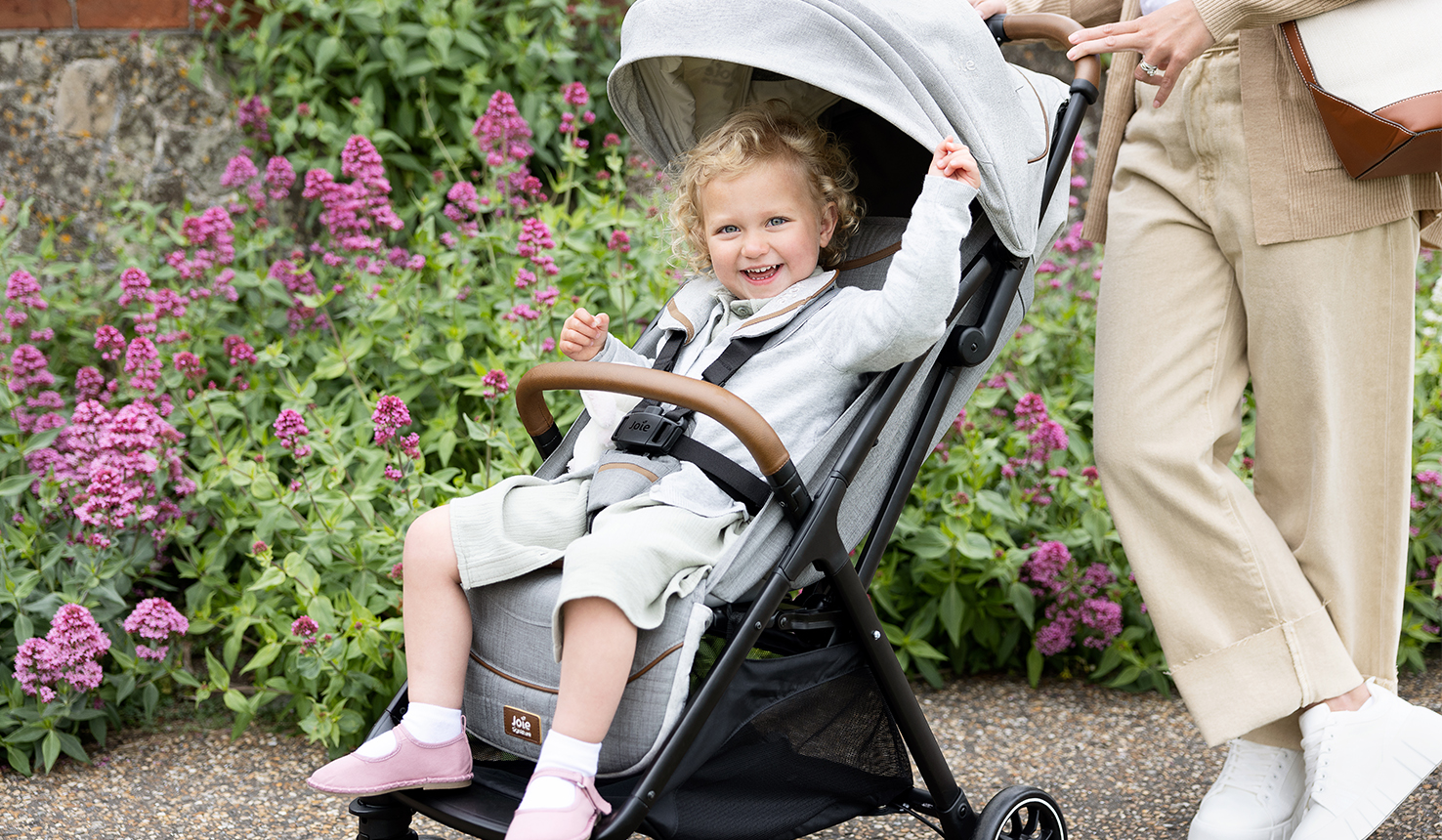 Ein blondes Mädchen sitzt im Joie Parcel Kinderwagen und lächelt.