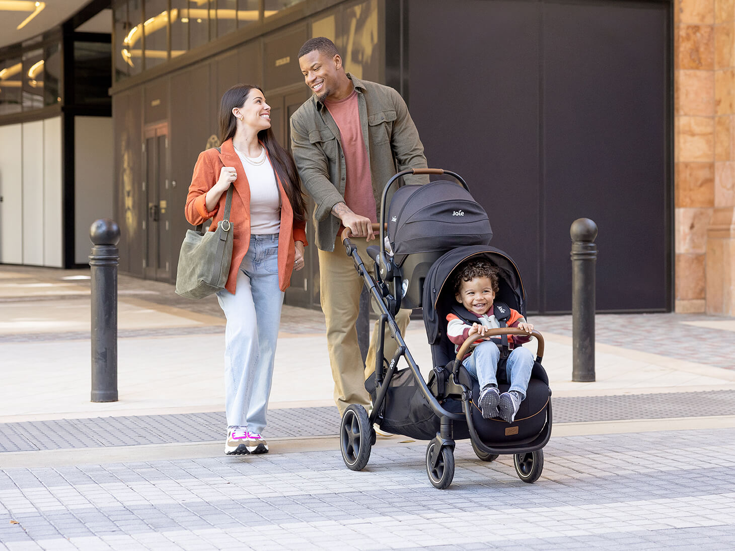 Mom and dad pushing infant and baby in chive stroller in shale with mint latch car seat and chive sibling seat
