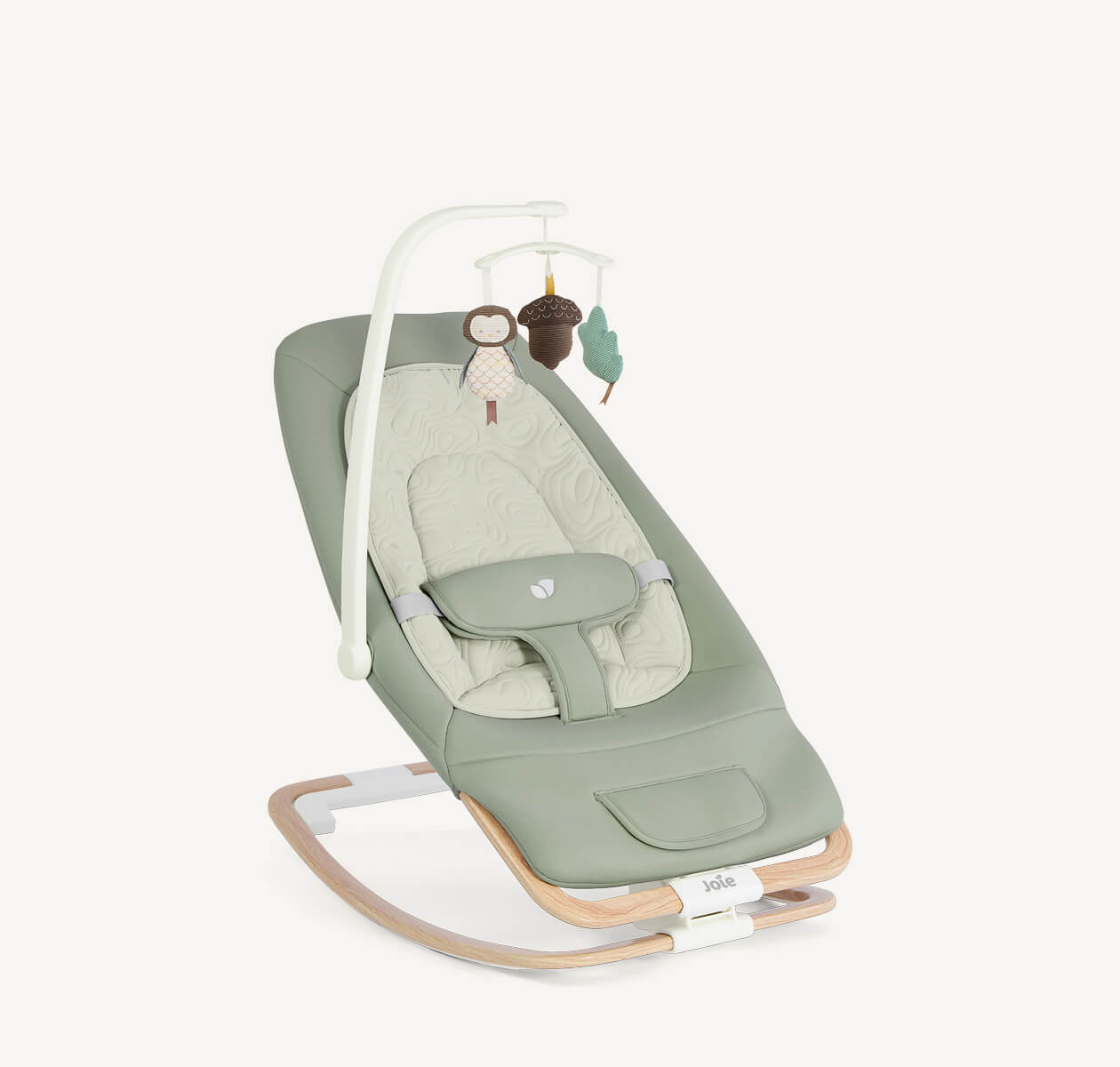 Joie Dreamer bouncer in light green with a light wooden base positioned at a right angle. The matching insert has a wavy stitching pattern and the attached mobile has 3 plush toys attached: an owl, acorn, and a green leaf.

