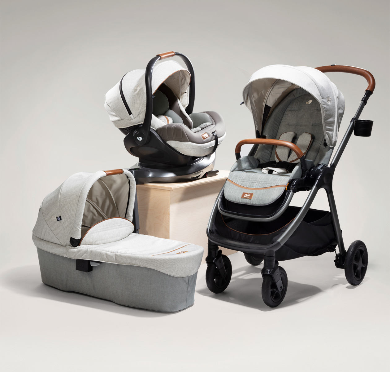 3 Joie Signature products in gray with brown leather accents: the Finiti pram, Ramble XL carry cot, and I-Level Recline infant car seat.