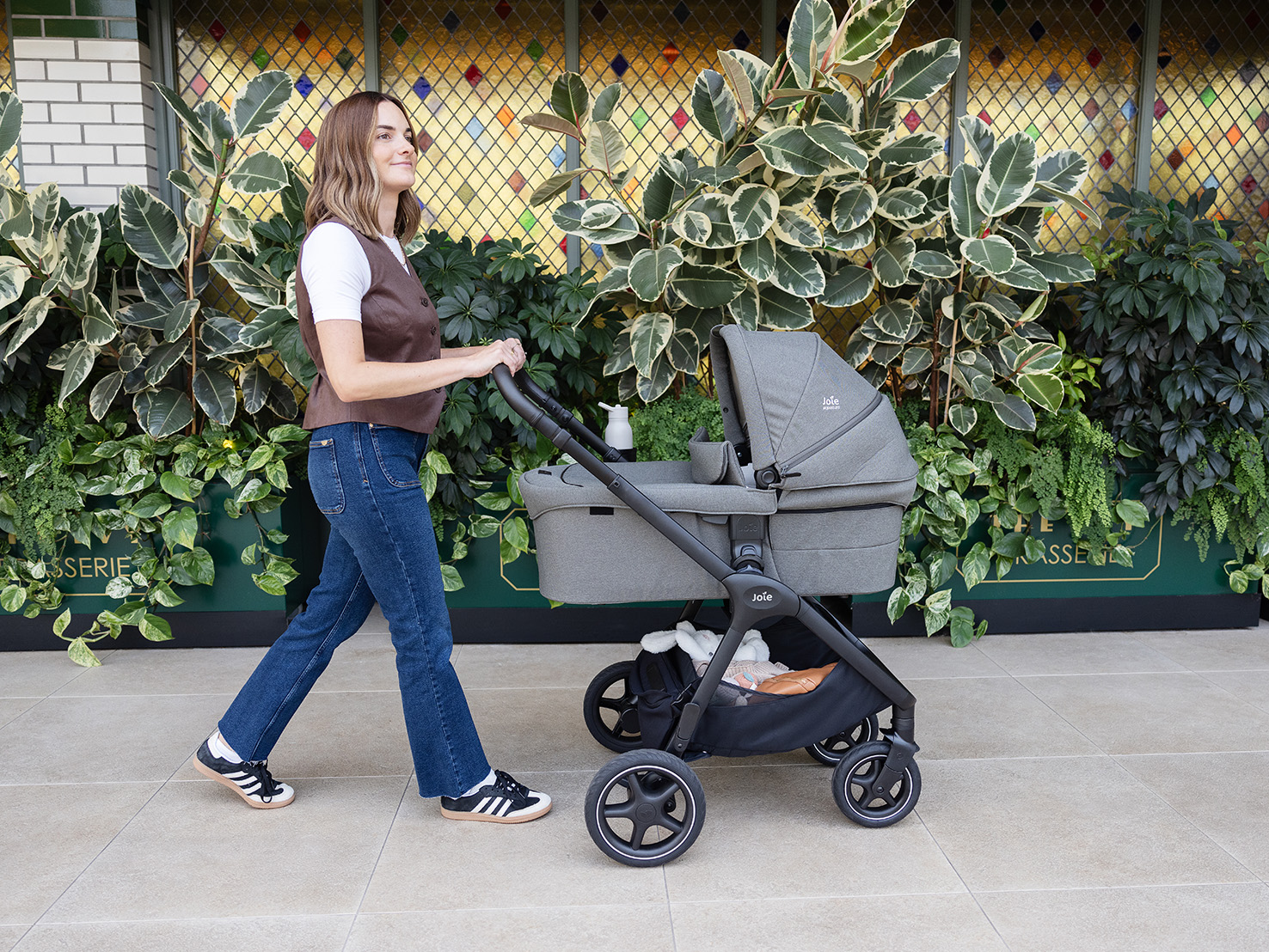 Mom strolls past a lush wall of greenery with her baby nestled in the cosy protection of the Joie Signature ramble xl carry cot.