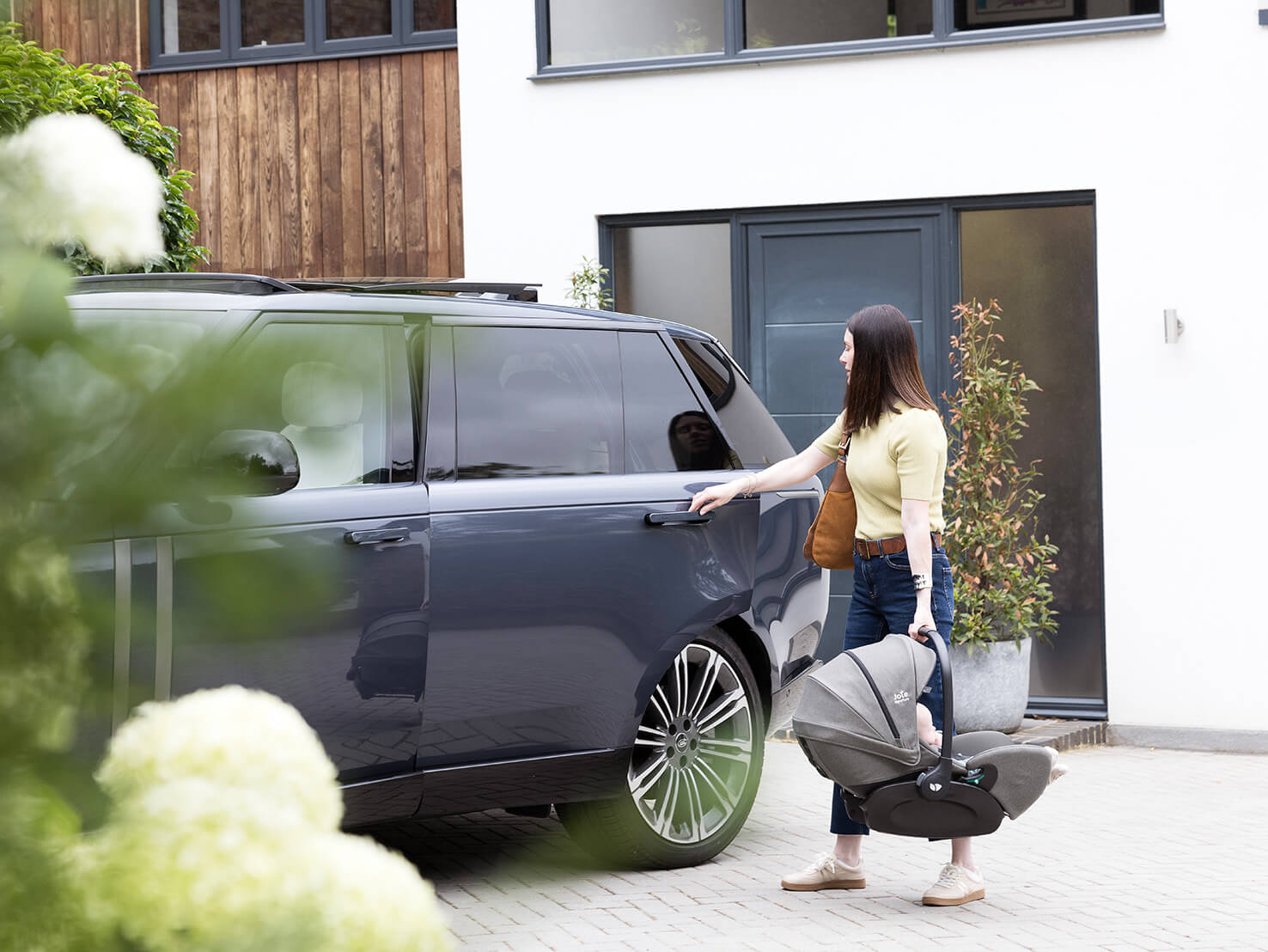 Mom carrying her newborn to the car in a Joie i-Level Pro deep reclining i-Size infant car seat. 
