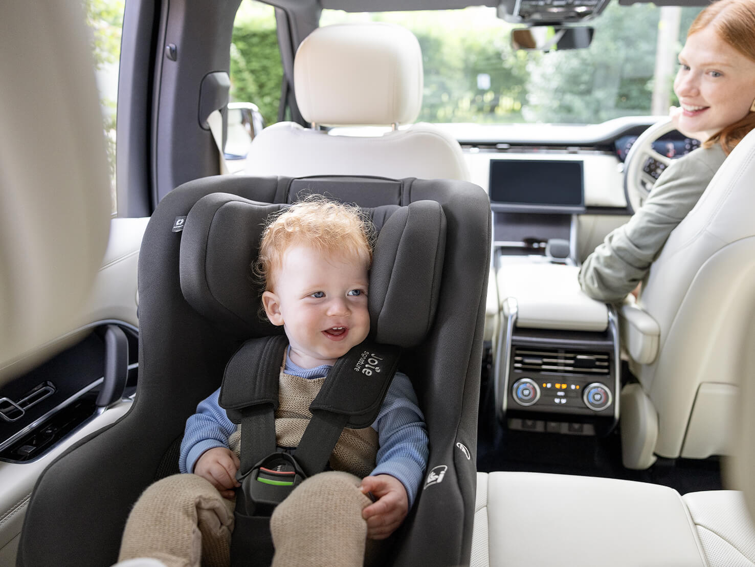 Young boy sitting rearward facing in the Joie Signature i-Prodigi Pro car seat while his mom is smiling back at him from the front seat with Swedish Plus Test, i-Size and R129 logos
