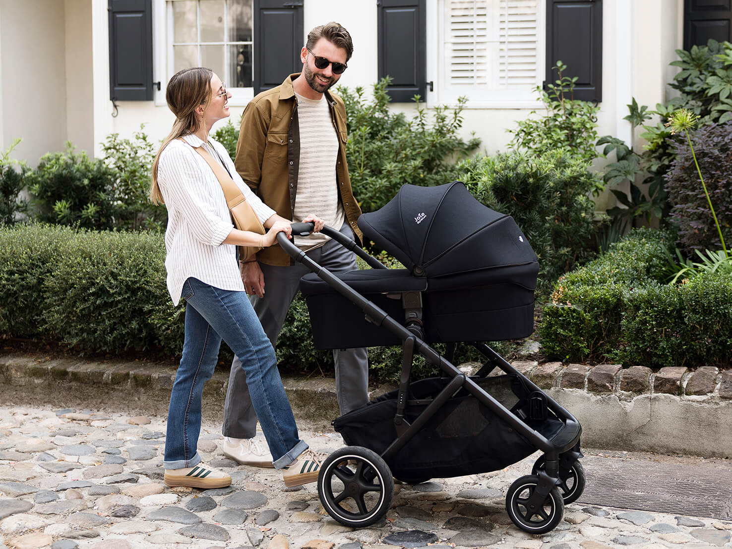 Mother and father pushing Joie Signature versiti pushchair in black, with ramble xl carry cot attached, along cobbled terrain.