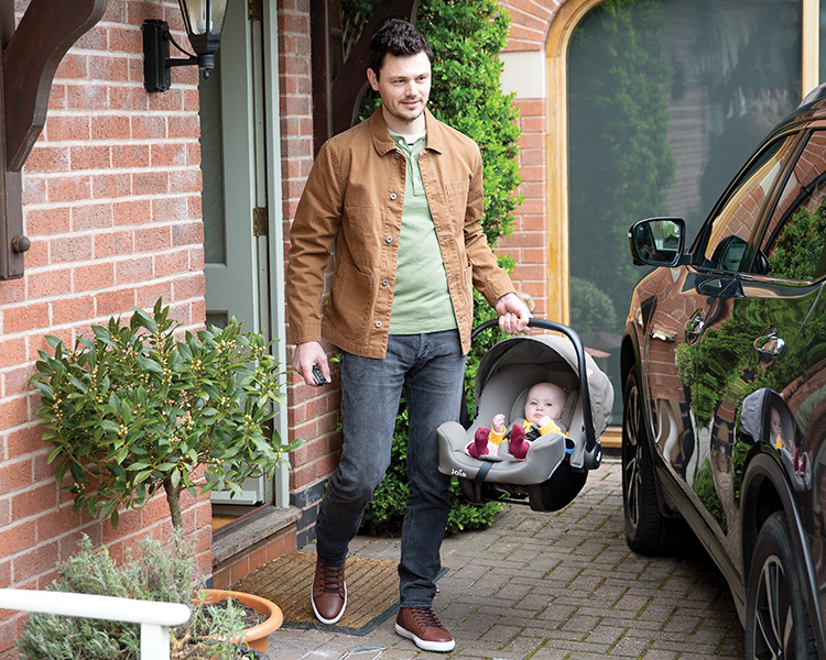 A man walks out of a brick house carrying a baby in a lightweight Joie i-Snug 2 infant car seat, heading toward a parked SUV. The baby is safely buckled in and looks comfortable, showcasing the ease of carrying and everyday convenience of the seat.
