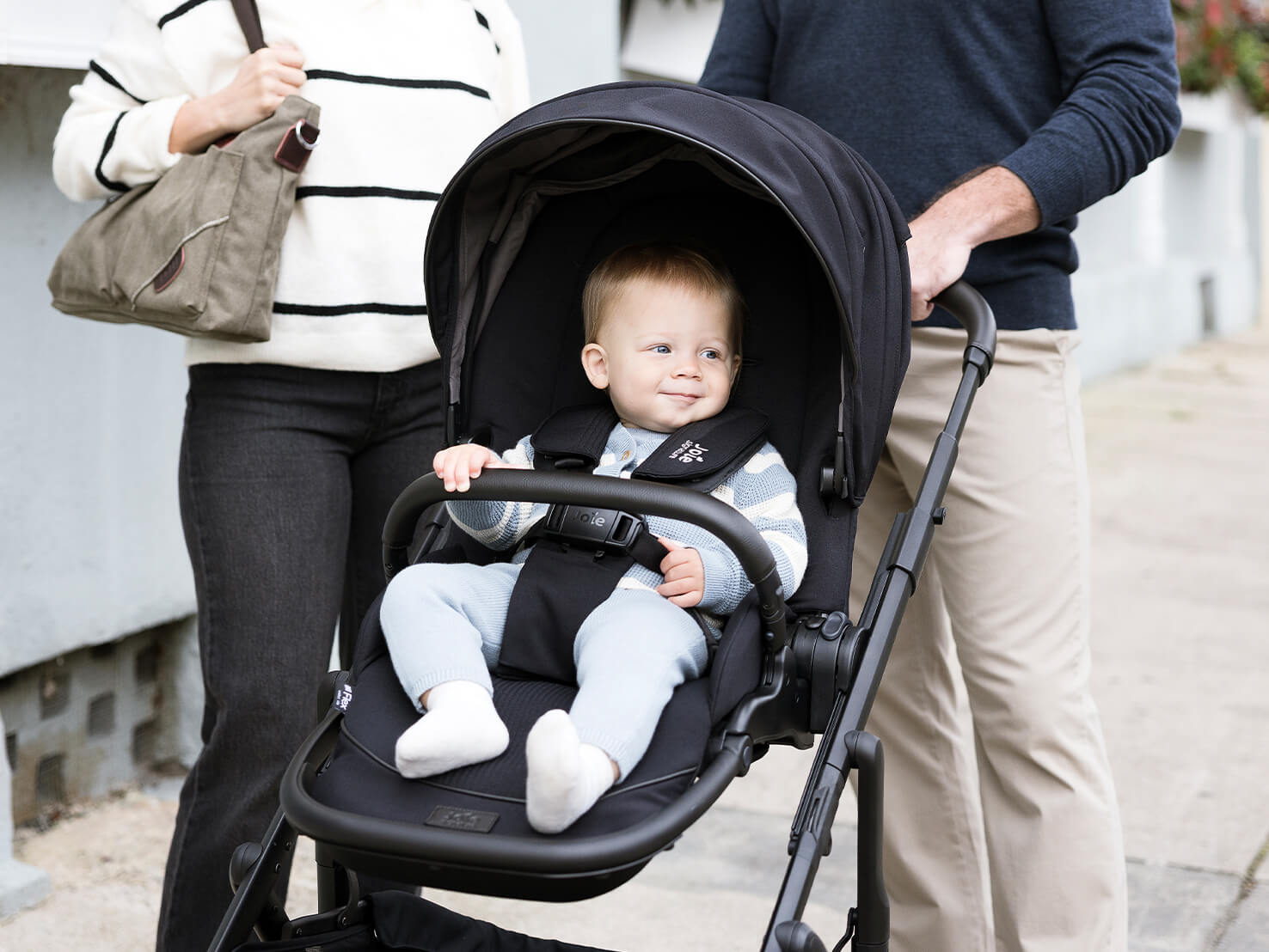 A mother and father pushing Joie Signature versiti pushchair in black with child in the seat, calf support is raised and UPF canopy is open.