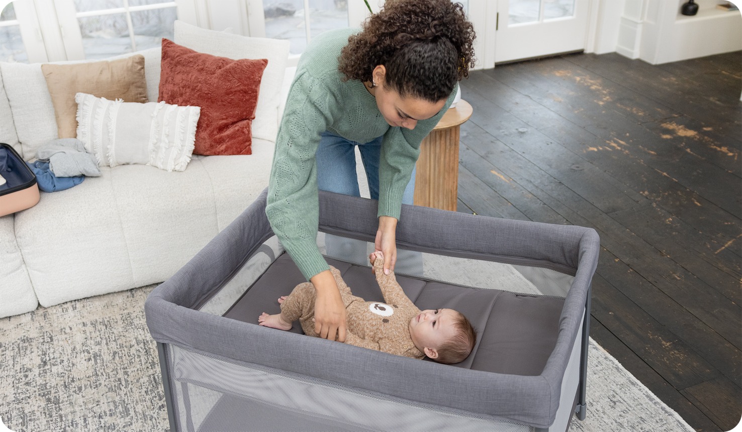 Mom playing with baby laying in amigo travel cot in bassinet mode
