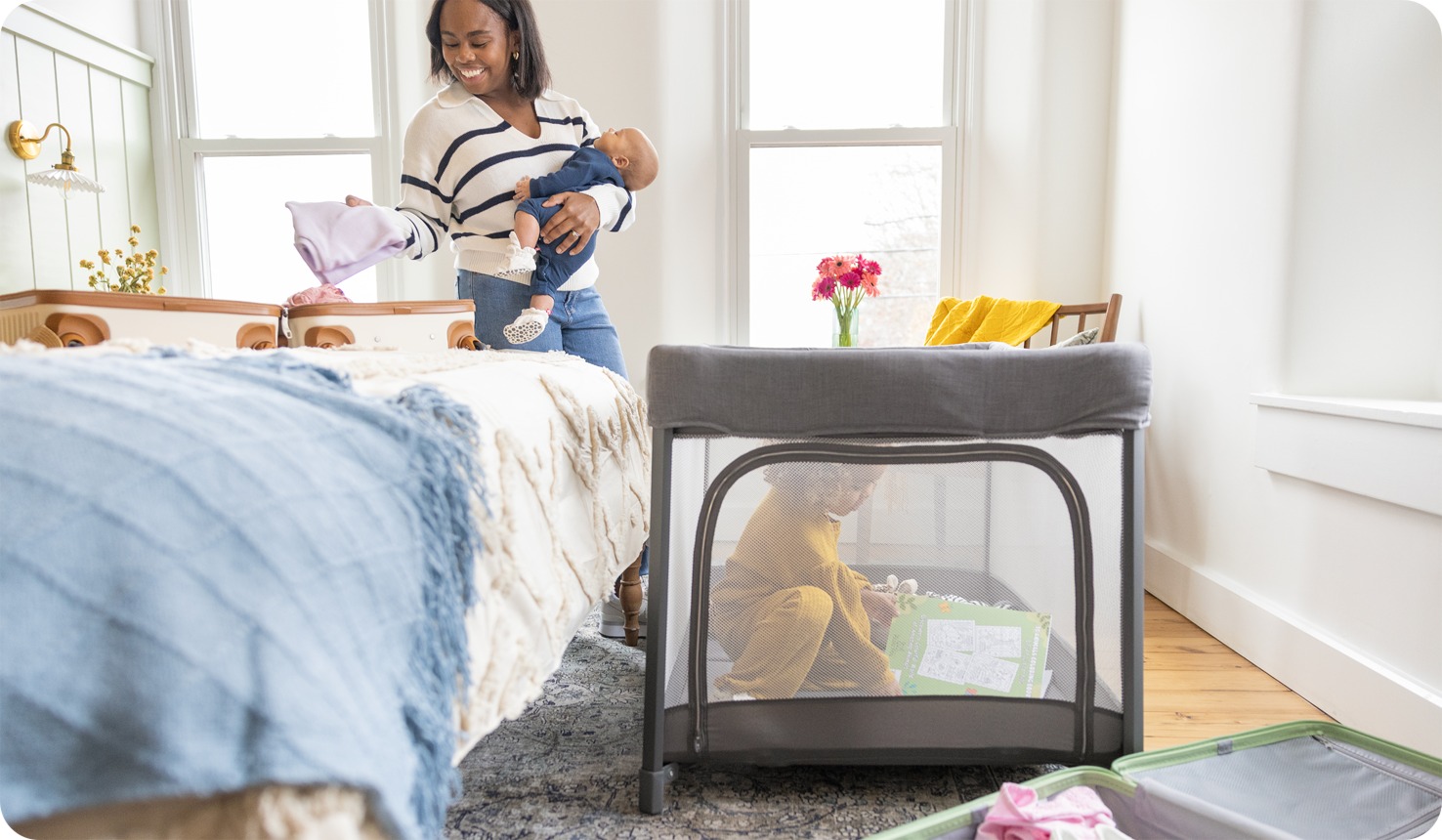 Mom holding baby while toddler plays next to her inside amigo travel cot in playard mode
