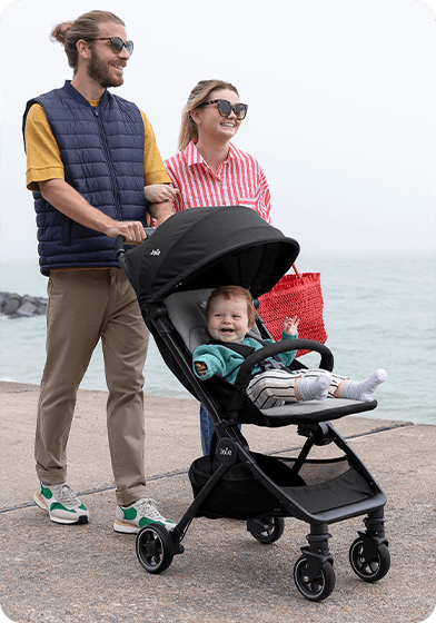 Mama und Papa gehen mit einem glücklichen Baby, das in einem schwarzen Joie Pact sitzt, spazieren. 