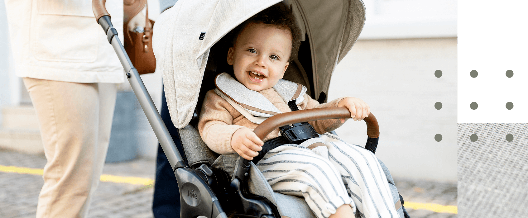 A young curly-haired toddler smiling toward the camera while being pushed in a gray Joie Finiti pram.