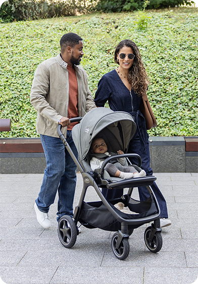 Couple strolling through a park setting, pushing the Joie Signature finiti pushchair in grey with baby comfortably riding in the world facing position.