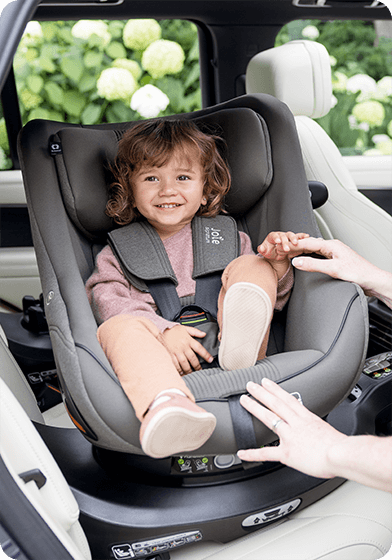 Tout-petit souriant installé dans le siège auto i-Harbour gris de Joie sur la base i-Base Encore, pendant que sa maman l’attache.