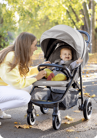 Matka se usmívá a klečí vedle miminka v kočárku Joie Litetrax Pro venku.