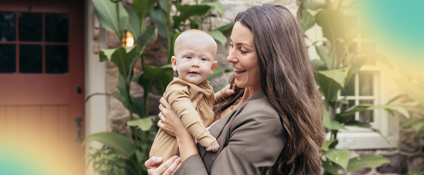 La madre sonr&iacute;e mientras sostiene a su beb&eacute; al aire libre, frente a una casa con plantas en el jard&iacute;n.