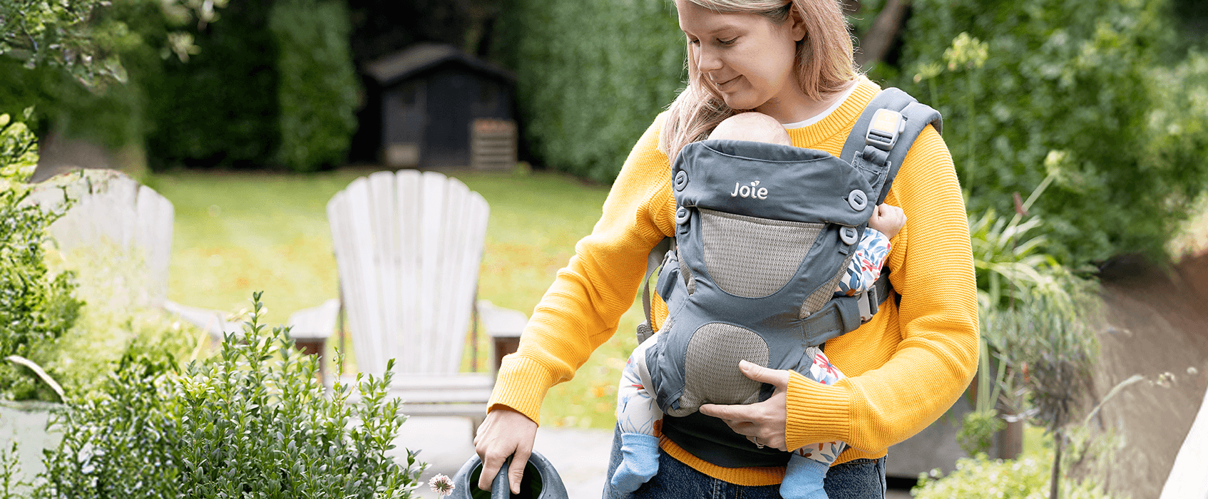 Parent wearing baby in the Joie Savvy ergonomic baby carrier while watering plants outdoors