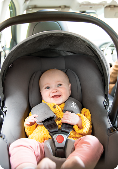 A happy baby in a yellow sweater is buckled into the lightweight Joie i-Snug 2 infant car seat inside a car.
