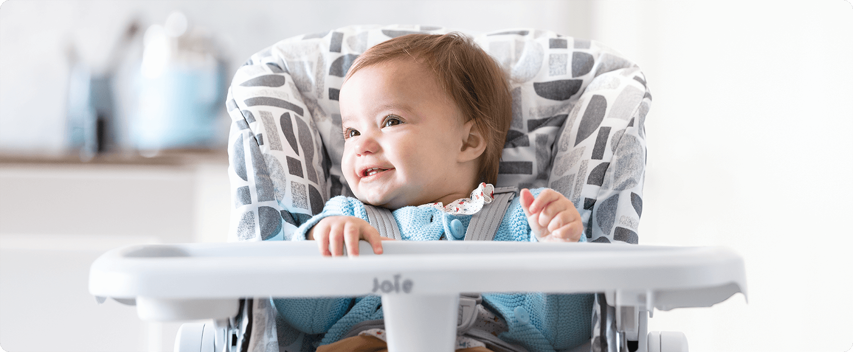 Das Baby sitzt in einem grauen und weißen 2-in-1-Hochstuhl Joie Snacker und schaut nach links.