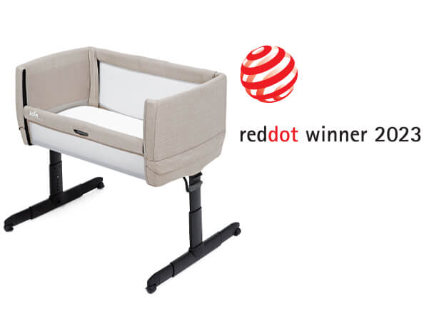 A tan Joie Roomie Go bedside crib at an angle facing to the left, with the Red Dot Design award logo above and to the right.