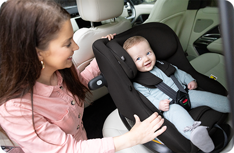 A smiling mother in a pink shirt buckles her baby into a black Joie i-Pivot™ 360 car seat installed in the back seat of a car.