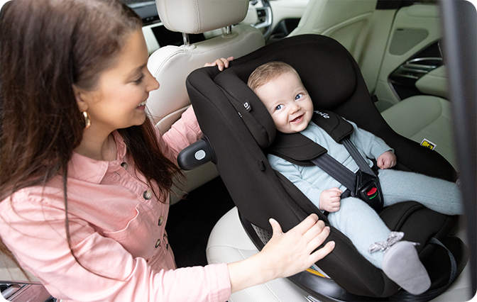 A smiling mother in a pink shirt buckles her baby into a black Joie i-Pivot™ 360 car seat installed in the back seat of a car.