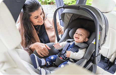 Smiling baby sitting in gray Joie i-Level Pro car seat while parent leans in with care.