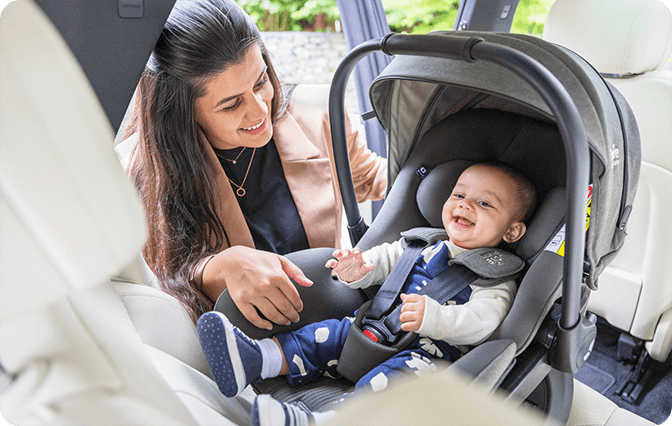 Smiling baby sitting in gray Joie i-Level Pro car seat while parent leans in with care.