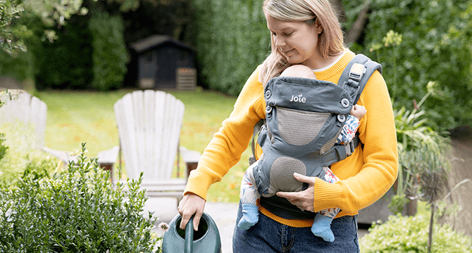 Parent wearing baby in the Joie Savvy ergonomic baby carrier while watering plants outdoors