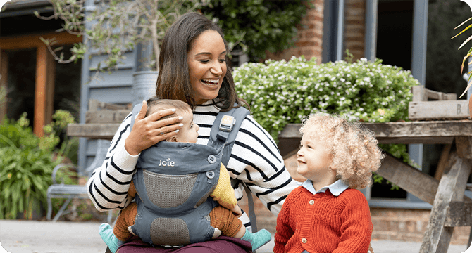 A mum and toddler sitting on a patio. Mum is wearing a baby in a blue Joie Savvy baby carrier.