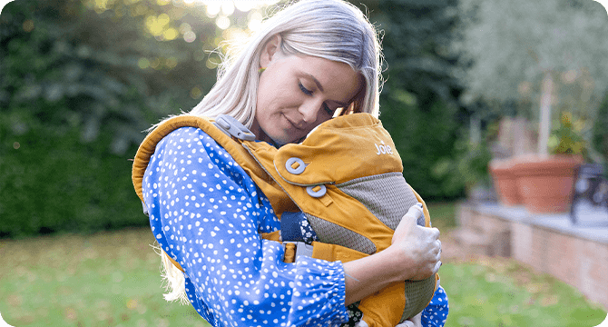 Mamá acurrucando a su bebé en un portabebés Joie Savvy amarillo mostaza