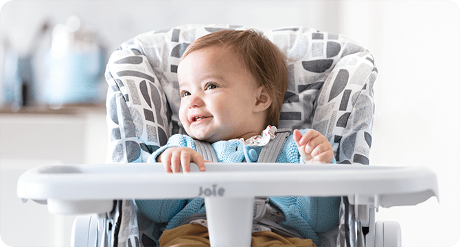 Baby sitting in a gray and white Joie Snacker 2in1 highchair, looking to the left.