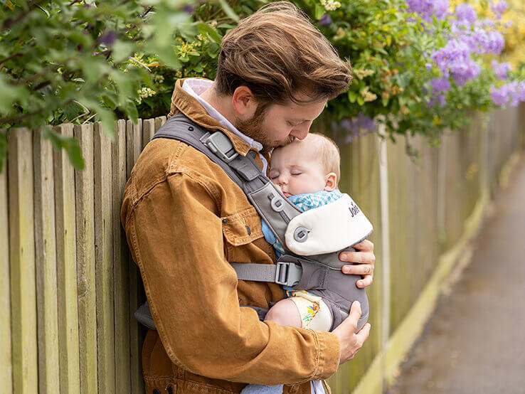 Dad wearing savvy baby carrier kissing baby's forehead as it sleeps soundly.