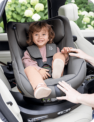 Tout-petit souriant installé dans le siège auto i-Harbour gris de Joie sur la base i-Base Encore, pendant que sa maman l’attache.