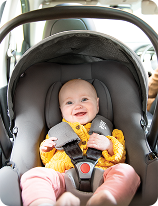 A happy baby in a yellow sweater is buckled into the lightweight Joie i-Snug 2 infant car seat inside a car.